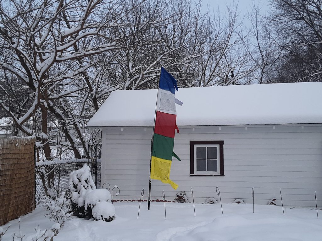 Tibeten prayer flags on a vertical pole (top to bottom: blue, white, red, green, yellow) blowing in the wind, in front of a garage, on snowy ground