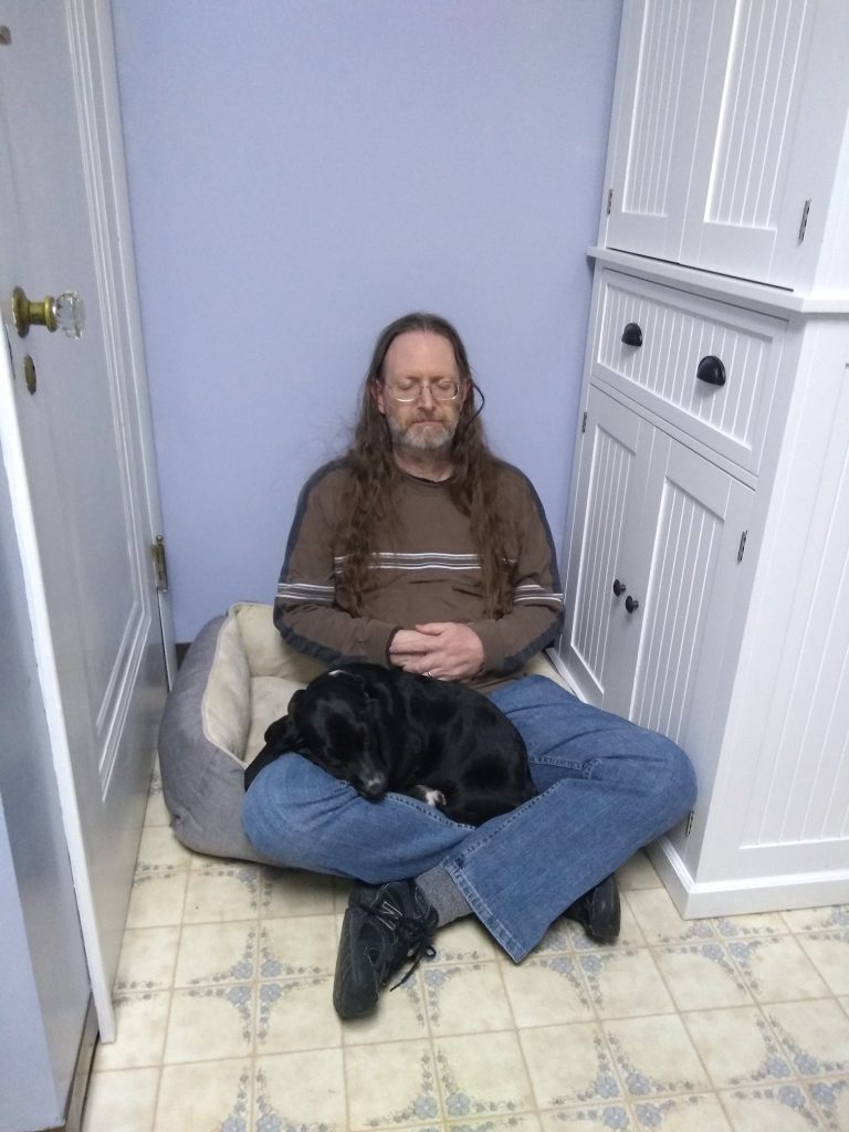 a long haired man with glasses is sitting, cross-legged, on a kitchen floor with a black basset hound puppy in his lap; both are asleep