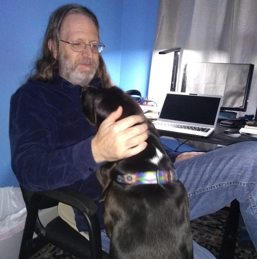 long haired, bearded man in blue room, sitting at a desk with an opened laptop on it, black basset hound standing on hind legs so the man can pet her